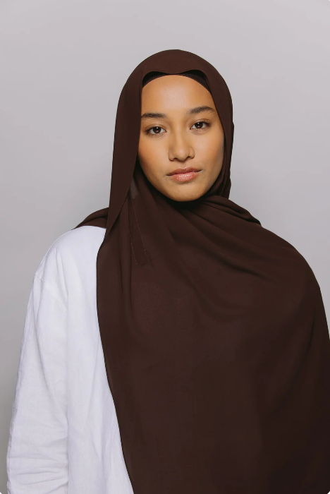 Woman wearing a brown hijab over a white shirt on a gray background